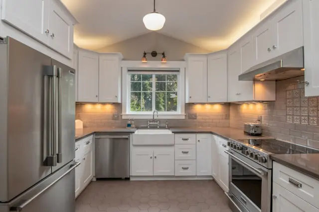 A modern kitchen with white cabinets, stainless steel appliances, a farmhouse sink, and a tiled backsplash, illuminated by under-cabinet lighting and a pendant lamp.