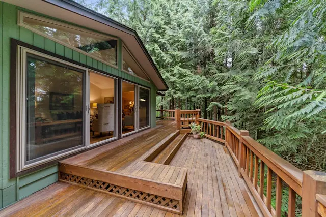 A wooden deck with a built-in bench extends from a green house with large sliding glass doors, surrounded by dense forest trees.