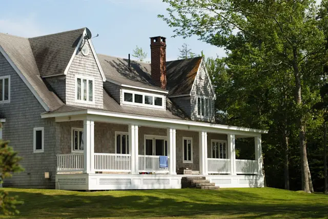A charming two-story house with a wraparound porch, gray shingle siding, and a brick chimney, set against a backdrop of lush green trees and lawn.