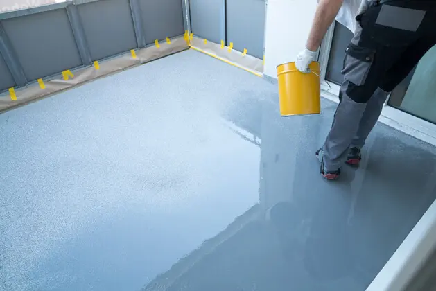 A worker in protective gear pours a glossy gray coating onto a freshly prepared floor, with yellow tape lining the edges.
