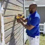 Professional painter in a blue uniform sanding a weathered wooden exterior wall with a yellow power sander, surrounded by a white fence and green lawn.