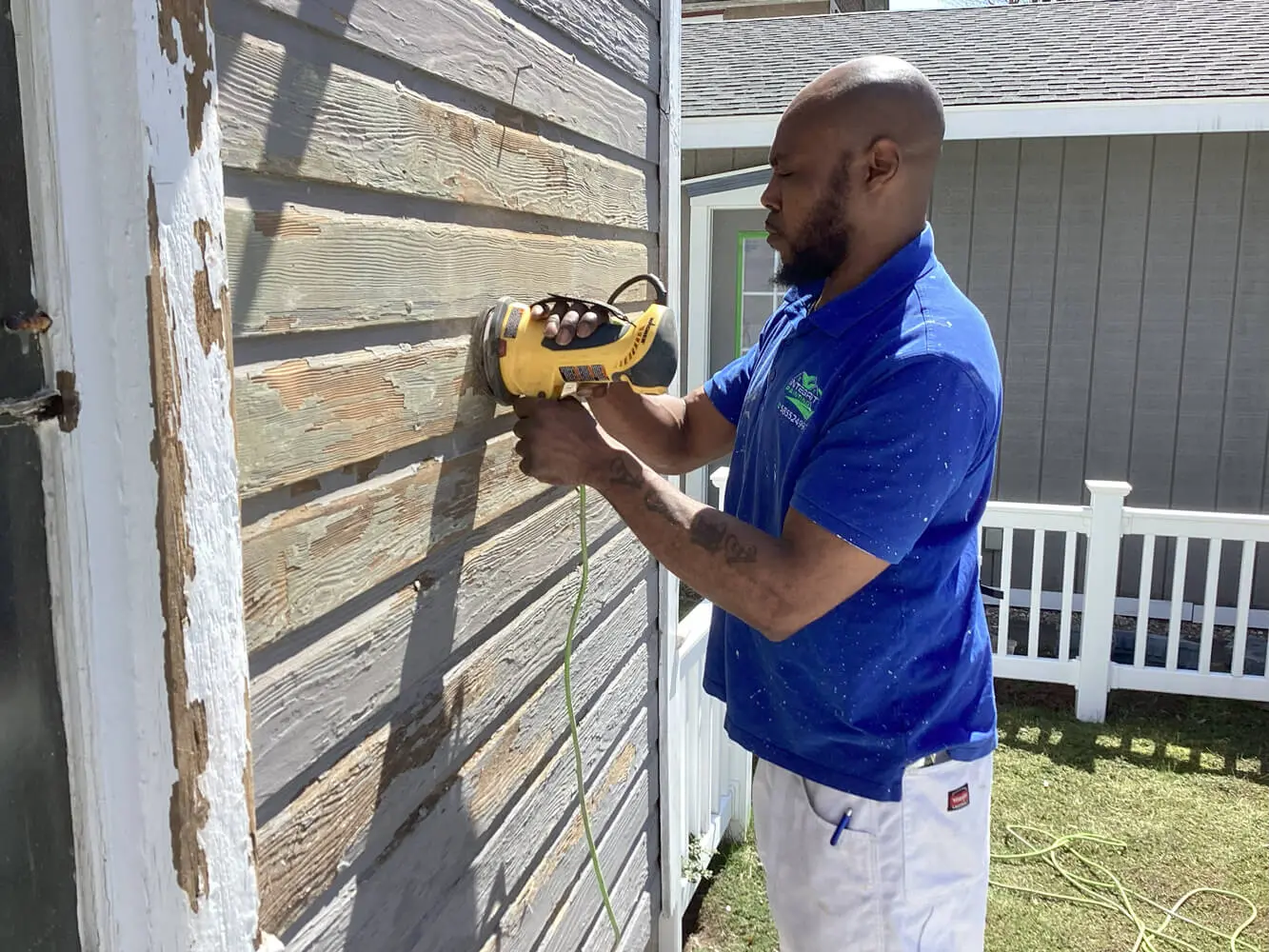 Professional painter in a blue uniform sanding a weathered wooden exterior wall with a yellow power sander, surrounded by a white fence and green lawn.