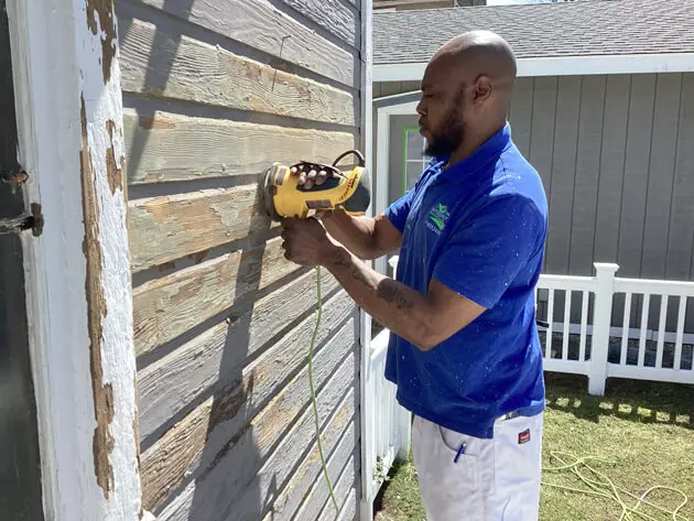 Professional painter in a blue uniform sanding a weathered wooden exterior wall with a yellow power sander, surrounded by a white fence and green lawn.