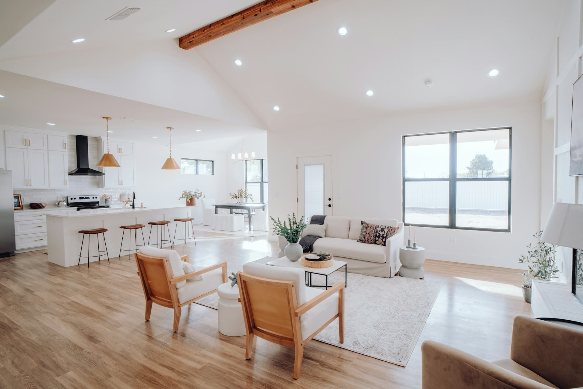 Modern open-plan living room with kitchen, wooden floors, white furniture, and large windows letting in natural light.