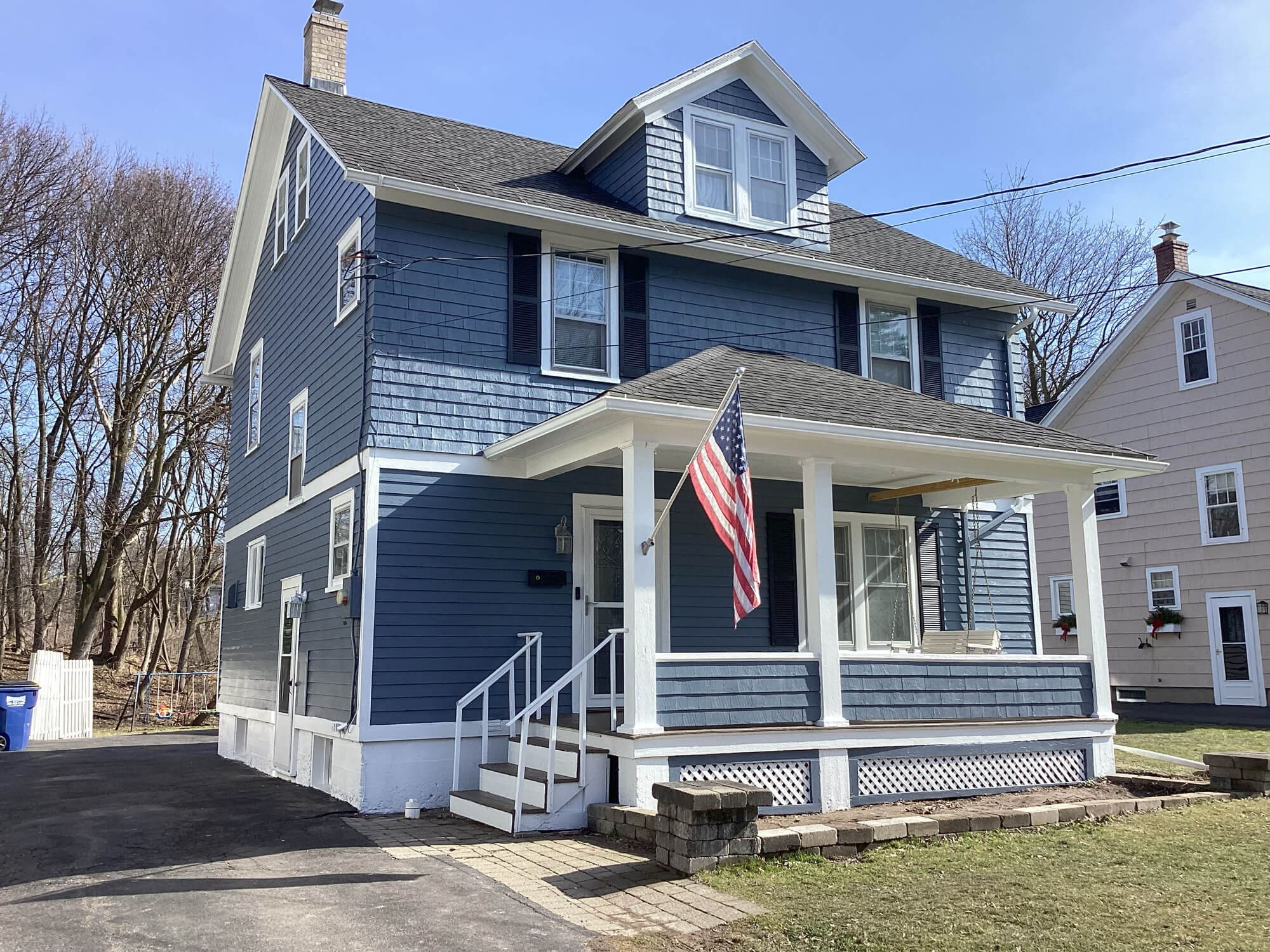 A charming blue two-story house with a white porch and American flag, surrounded by trees, ideal for real estate and home decor