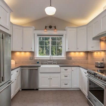 A modern kitchen with white cabinets, stainless steel appliances, a farmhouse sink, and a tiled backsplash, illuminated by under-cabinet lighting and a pendant lamp.