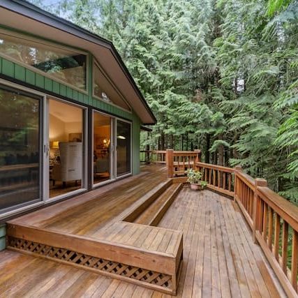 A wooden deck with a built-in bench extends from a green house with large sliding glass doors, surrounded by dense forest trees.