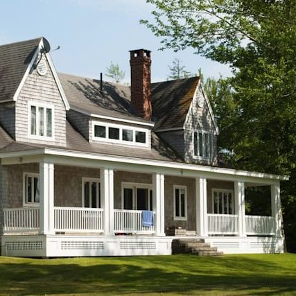A charming two-story house with a wraparound porch, gray shingle siding, and a brick chimney, set against a backdrop of lush green trees and lawn.