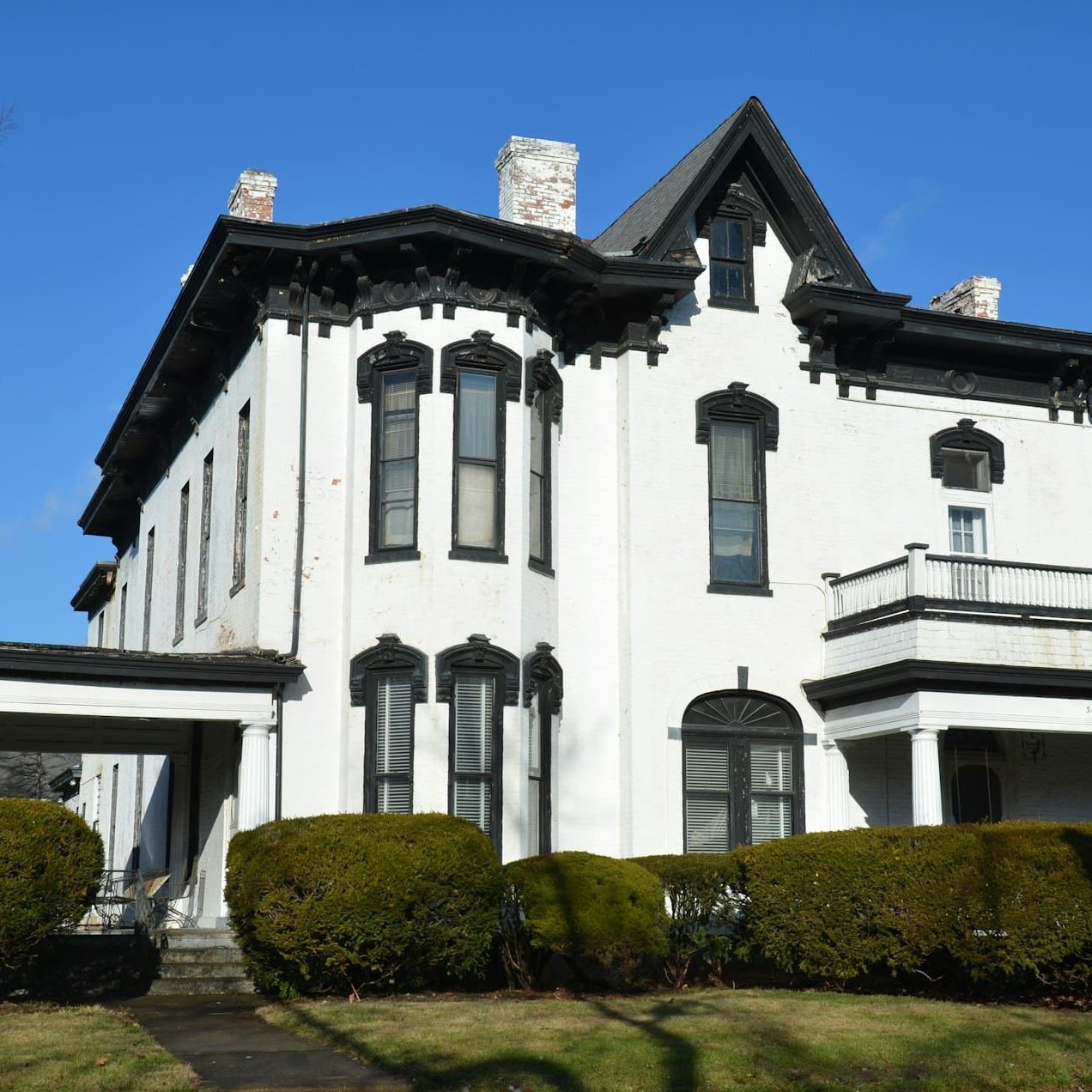 Historic white house with black trim, columns, and balcony, surrounded by greenery under a clear blue sky.