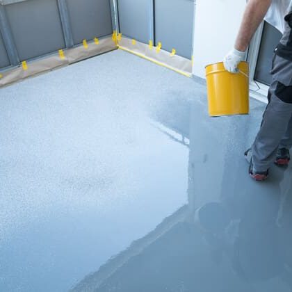 A worker in protective gear pours a glossy gray coating onto a freshly prepared floor, with yellow tape lining the edges.