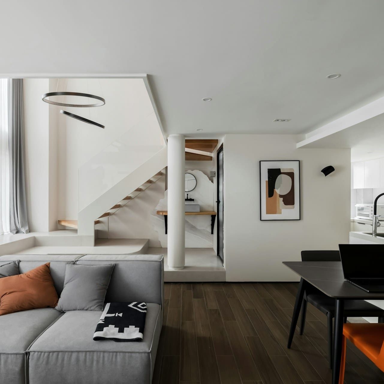 Modern minimalist living room with gray sofa, orange chair, wooden floor, and open kitchen with staircase.