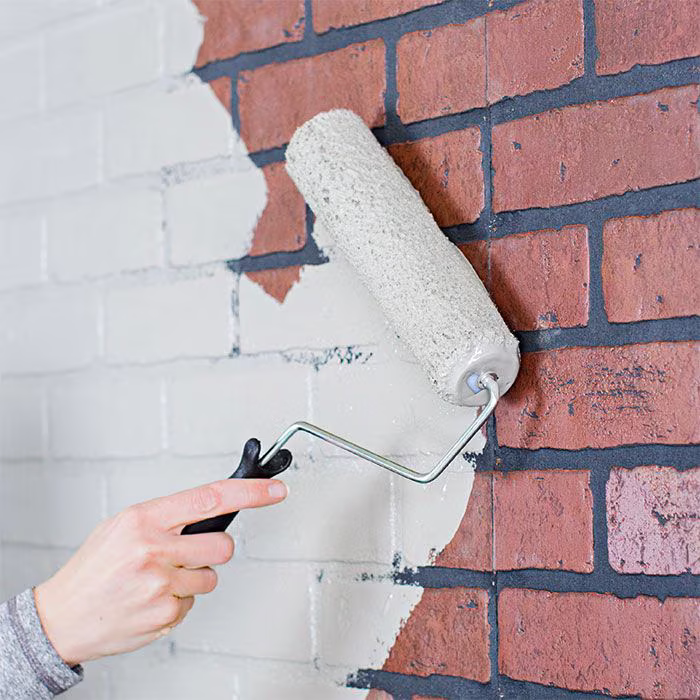 A hand using a paint roller to apply white paint over a brick wall.